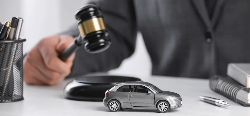 A lawyer in a gray business suit from Illinois Lawyers 365 sits behind a white desk in the blurred background gripping a black and gold judge's gavel and bringing it down toward its black circular sound block while in the sharp foreground a detailed silver die-cast toy sedan sits centered on the desk surface with a black mesh pen holder filled with writing instruments visible on the left edge of the frame and a stacked book and silver pen on the right — a legal concept image representing the courtroom authority an attorney carries when advocating for a client's rights following a car accident Illinois.