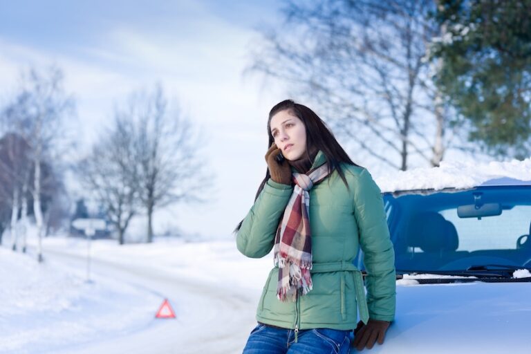 A young woman with long dark hair wearing a mint green puffer jacket- a plaid scarf in red and tan tones- and brown leather gloves leans against the snow-covered hood of a blue vehicle on the shoulder of a snow-blanketed rural road pressing a phone to her ear with a worried and distant expression on her face with a red triangular emergency warning reflector placed on the road surface behind her and a row of bare snow-dusted trees stretching along both sides of the empty white road into the pale winter sky — depicting the stressful moment a driver calls for help immediately following a winter weather car accident in Illinois.