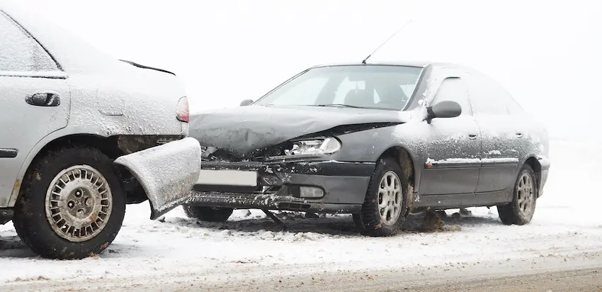 A dark charcoal gray sedan coated in a thin layer of snow and road grime has its entire front hood crumpled downward and the bumper completely detached and hanging off where it has driven into the rear end of a silver vehicle on an icy snow-covered road with both vehicles dusted in white and the surrounding environment completely white from heavy snowfall or blizzard conditions with minimal visibility in the distance — illustrating the dangerous road conditions that contribute to winter weather car accident collisions and could use help from Illinois Lawyers 365.