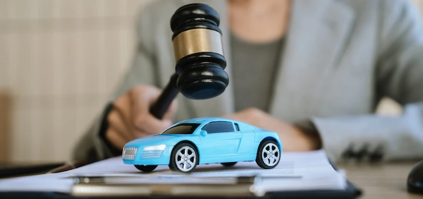 An attorney from Illinois Lawyers 365 in a gray blazer holds a gavel over a small blue toy car resting on legal documents at a desk- visually representing the judicial authority and legal process involved in resolving a car accident case through negotiation or litigation in Illinois.