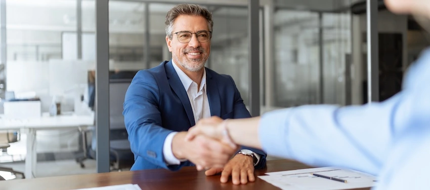A confident middle-aged attorney in a navy blazer and glasses smiles while shaking hands with a client across a desk with documents in view- representing the successful resolution of a car accident case in which the victim received a favorable settlement in Illinois.
